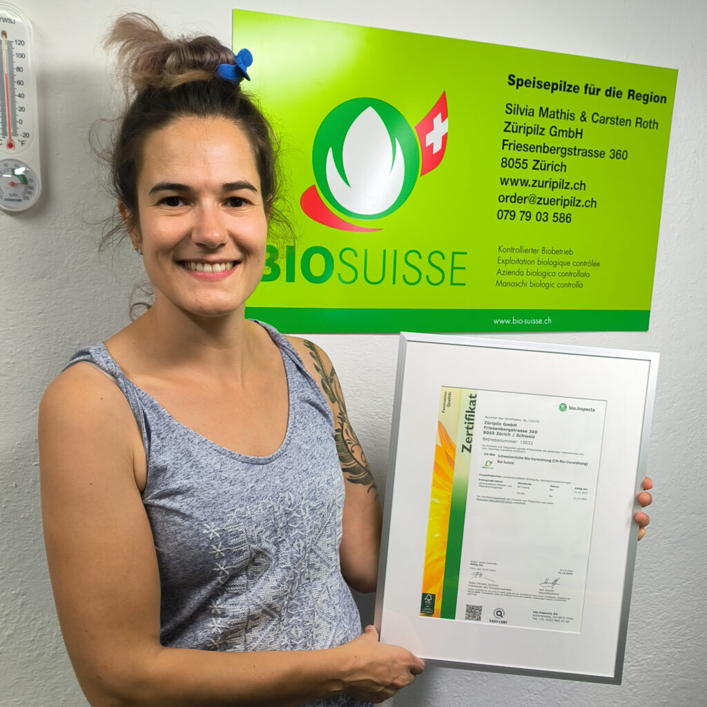 Mehr als ein Siegel: Unser Bekenntnis zu höchster Qualität und Werten Silvia steht lächelnd vor einem Bio Suisse Plakat der Firma Züripilz und hält stolz das Bio Suisse Zertifikat in den Händen.